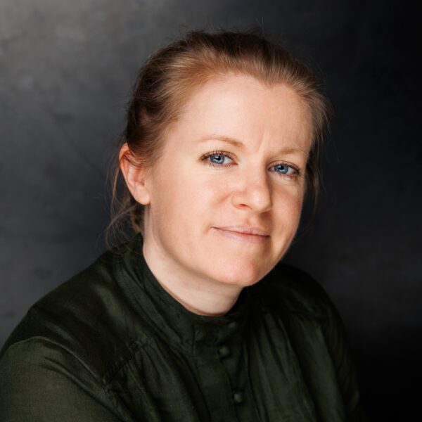 A woman with light skin, blue eyes, and light brown hair pulled back, wearing a dark green top, sits in front of a dark, neutral background and looks at the camera with a slight smile.