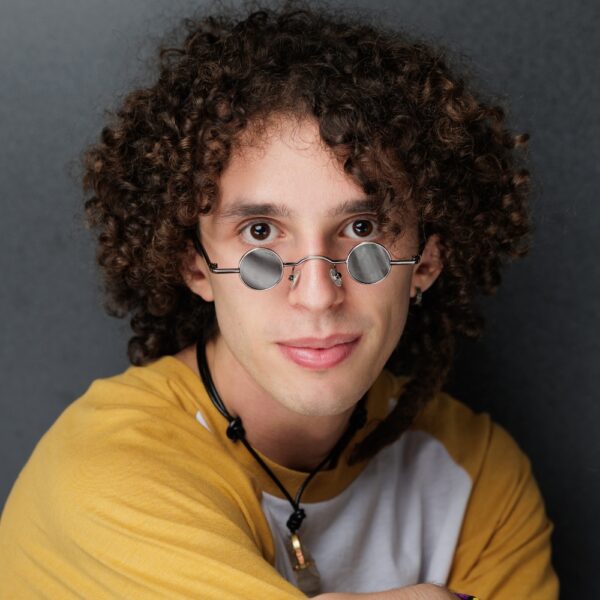 A young person with curly brown hair, wearing round, reflective glasses, a yellow and white shirt, and necklaces, sits against a dark background, looking at the camera with a slight smile.