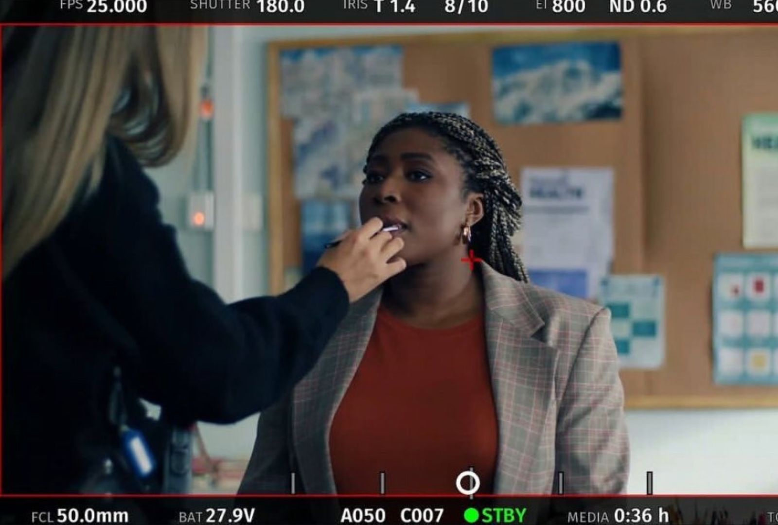 A woman in a red shirt and blazer is having her make-up done by a make-up artist on the set of Heartbreak High.
