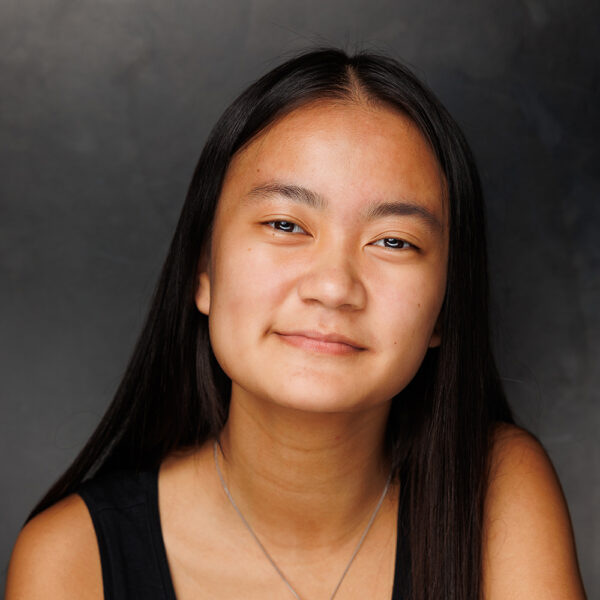 A young person with long, straight dark hair and a slight smile looks at the camera, wearing a black top and a silver necklace, against a dark grey background.