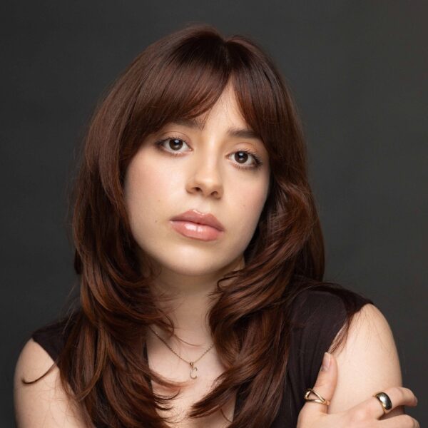 A young woman with medium-length brown hair and fringe poses against a dark background, wearing a black top and gold rings, with a calm and neutral expression.