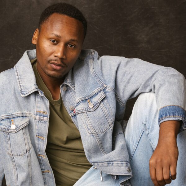 A man with short hair wearing a light blue denim jacket and jeans over a green shirt sits casually against a dark, textured background, with his arm resting on his knee and a relaxed expression.