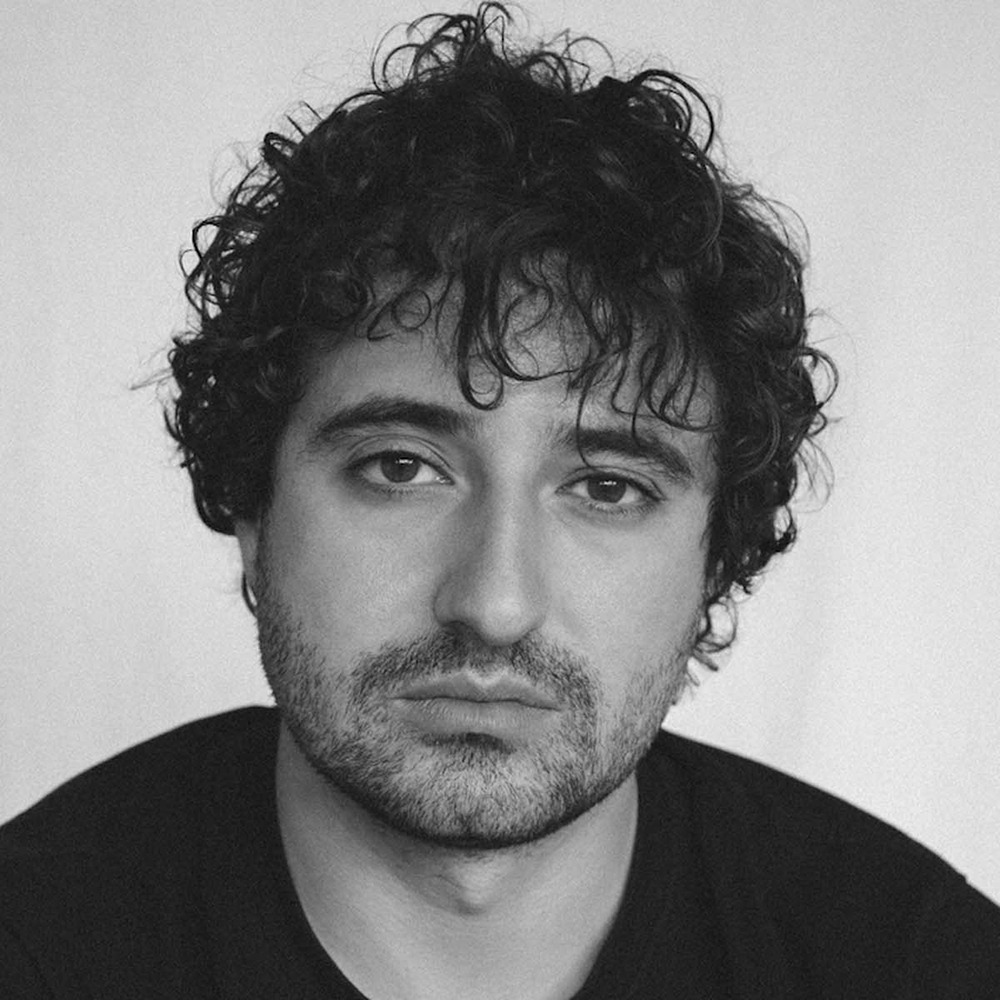A man with curly dark hair and a short beard looks directly at the camera with a neutral expression. He is wearing a dark shirt and is photographed against a plain, light background.