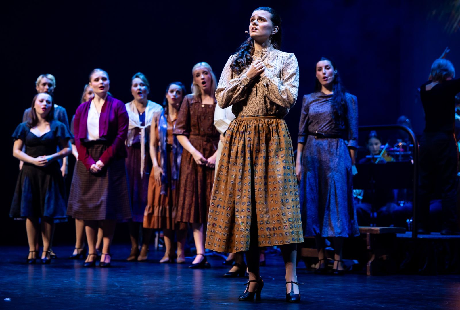 A woman in a long skirt on stage singing with a chorus of performers behind her.
