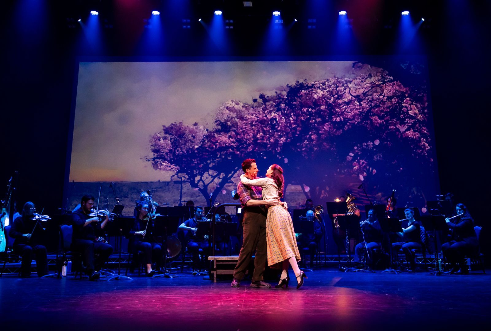 Two actors embrace on stage with musicians behind them and a tree projected onto a screen.