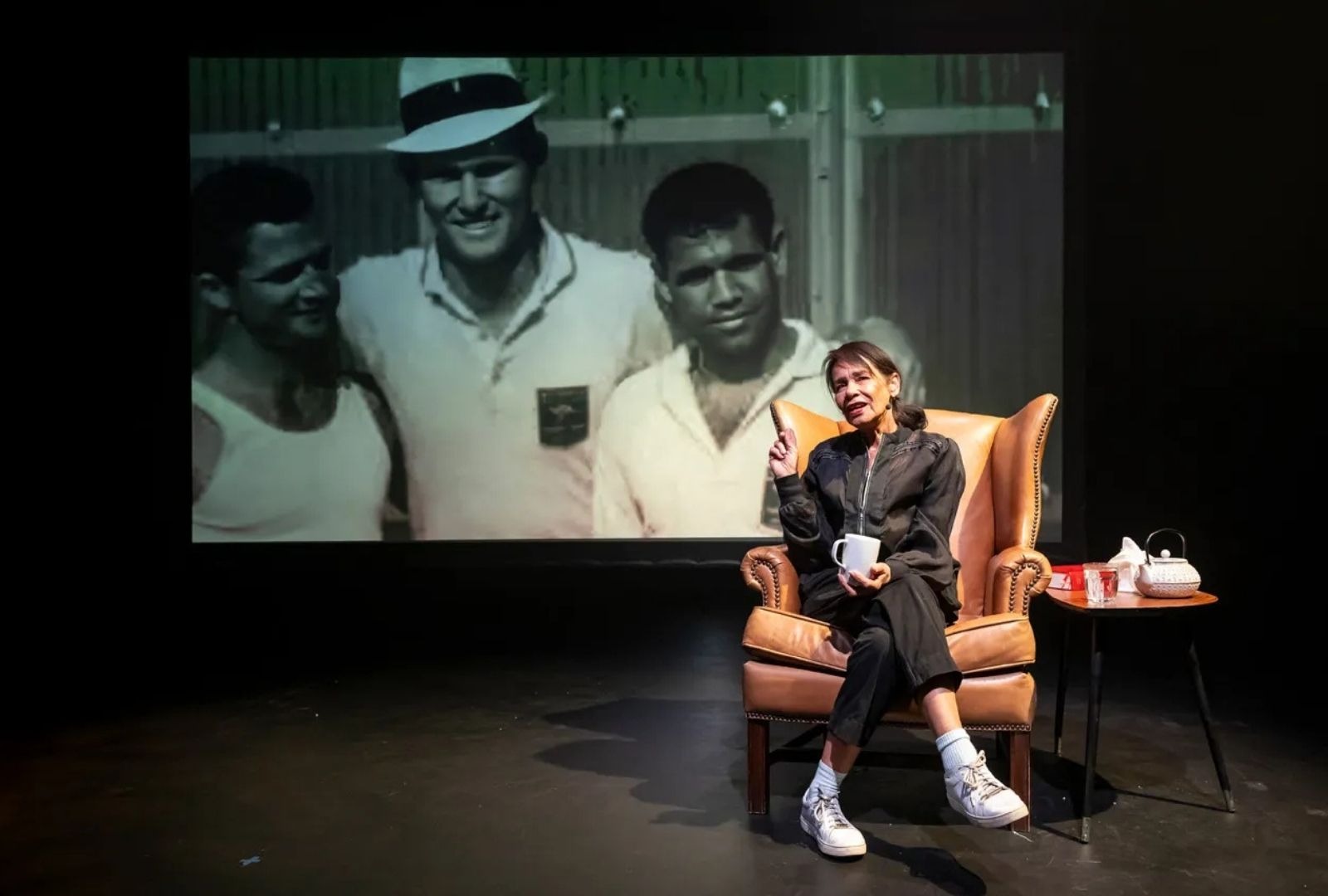 A woman sitting on a chair with a projection of three men behind her. She is telling a story.