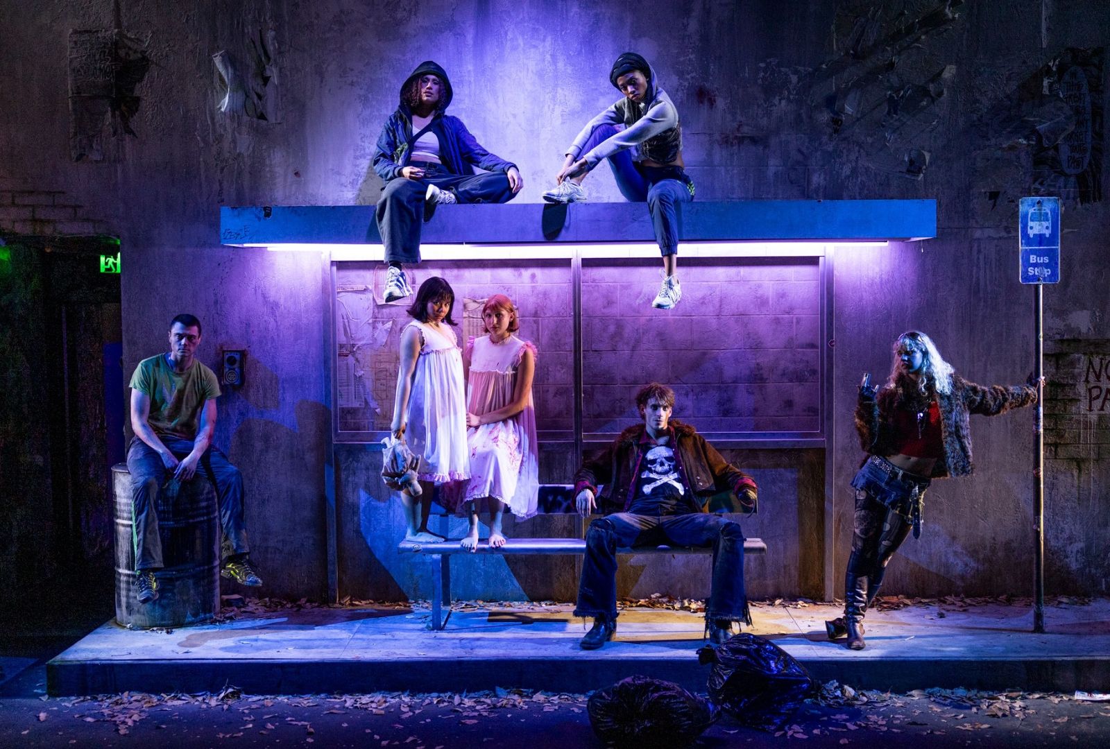 Seven people pose dramatically under a purple-lit bus stop, evoking a moody, theatrical atmosphere. They wear casual and edgy clothing.
