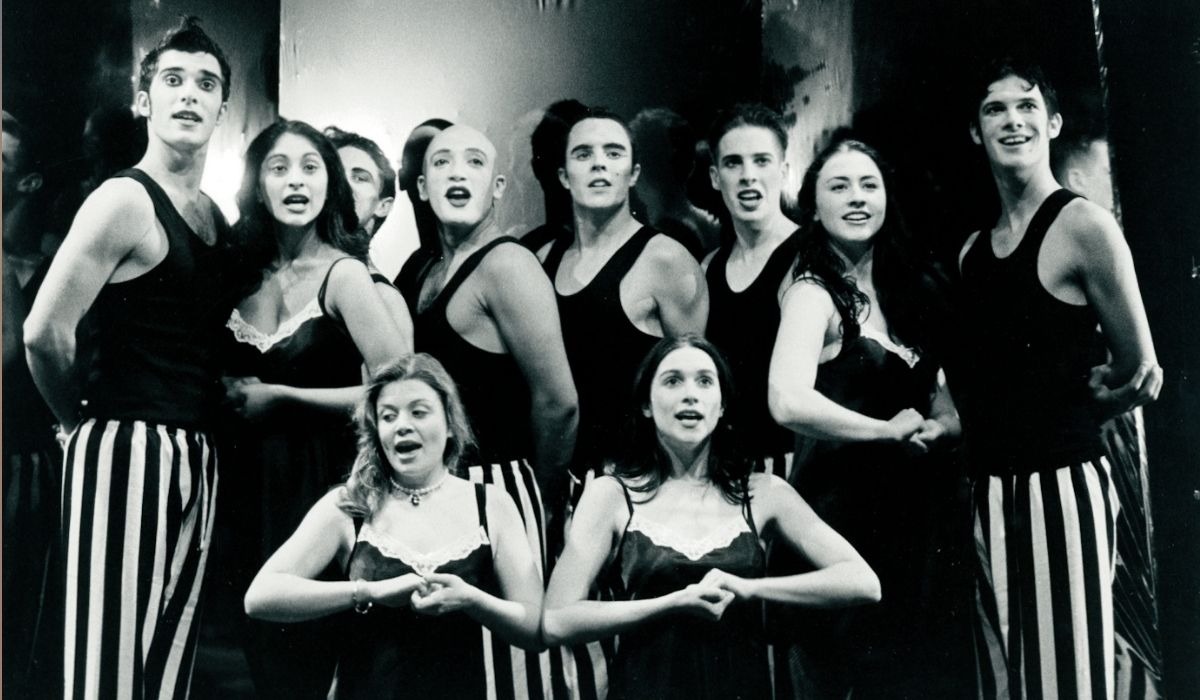 A group of nine performers on stage, wearing black tops and striped pants, expressively singing together. The mood is energetic and theatrical.