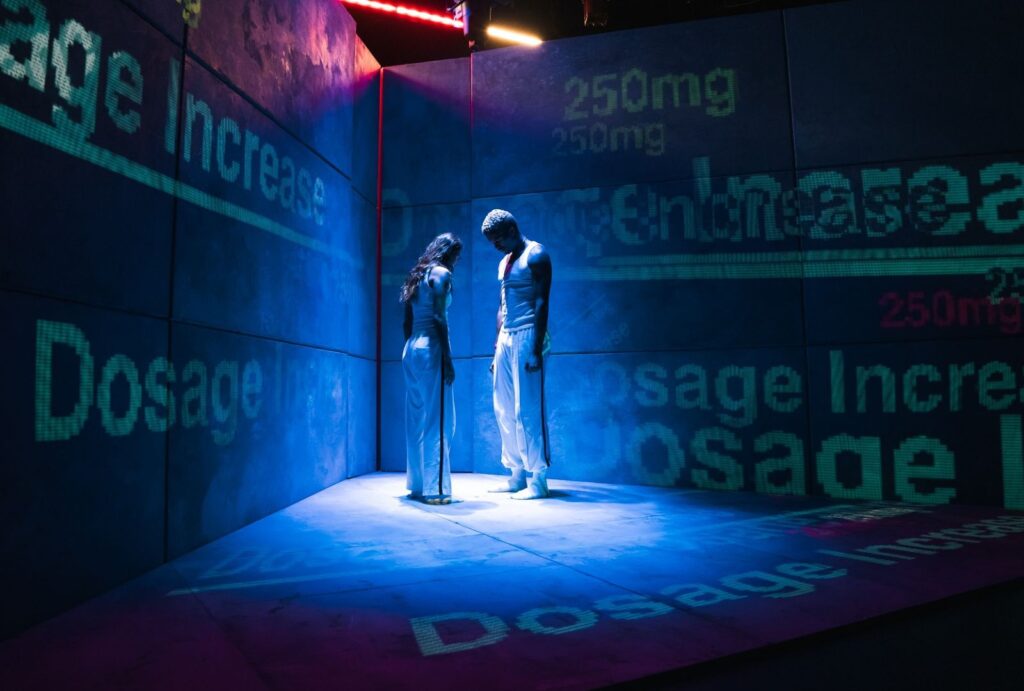Two people stand under blue and pink lights in a corner of a room, both looking down. The walls and floor display large projected words like Dosage Increase and 250mg.