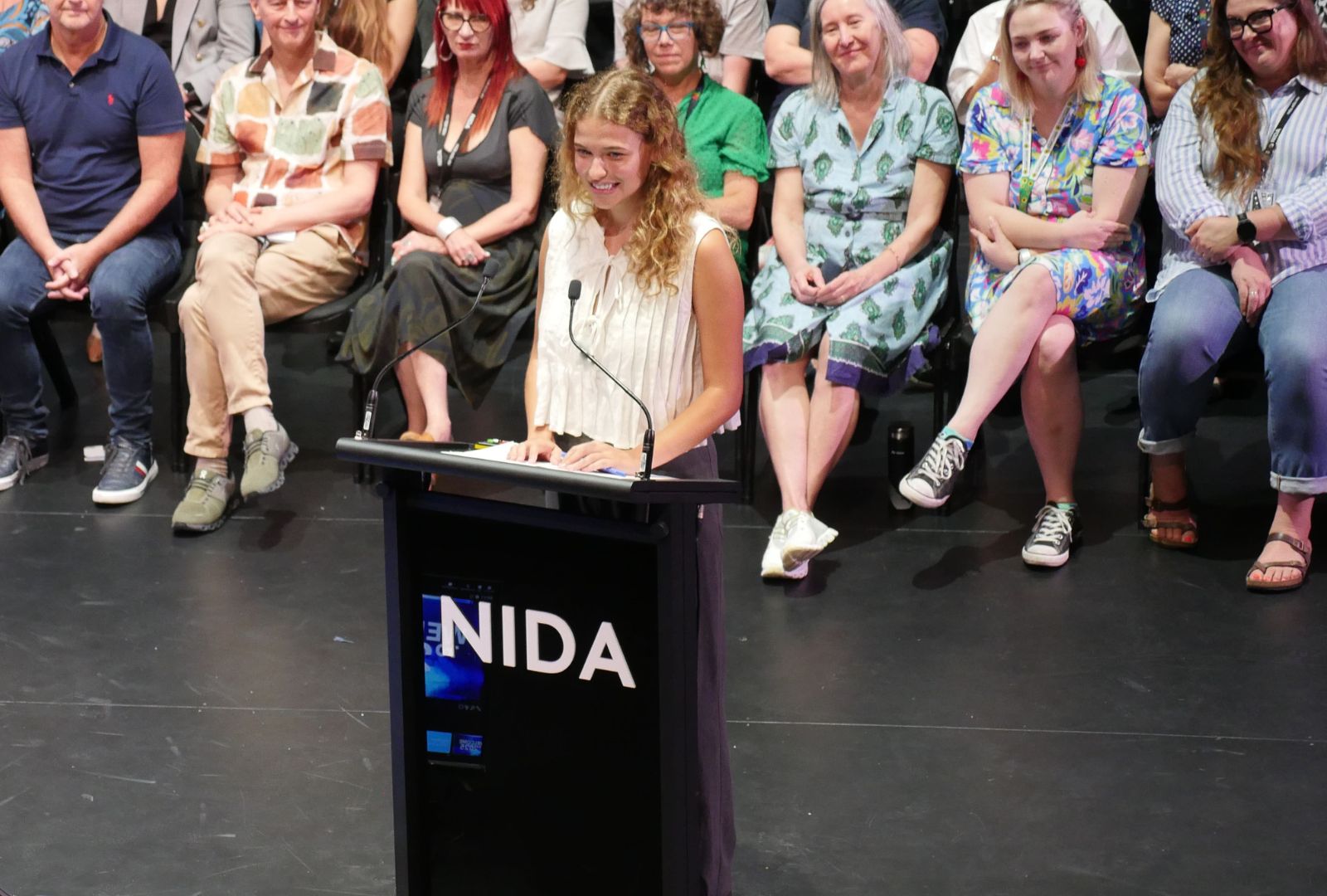Clare Hughes on stage at Open Day standing at a podium that reads 'NIDA' with members of staff behind her.