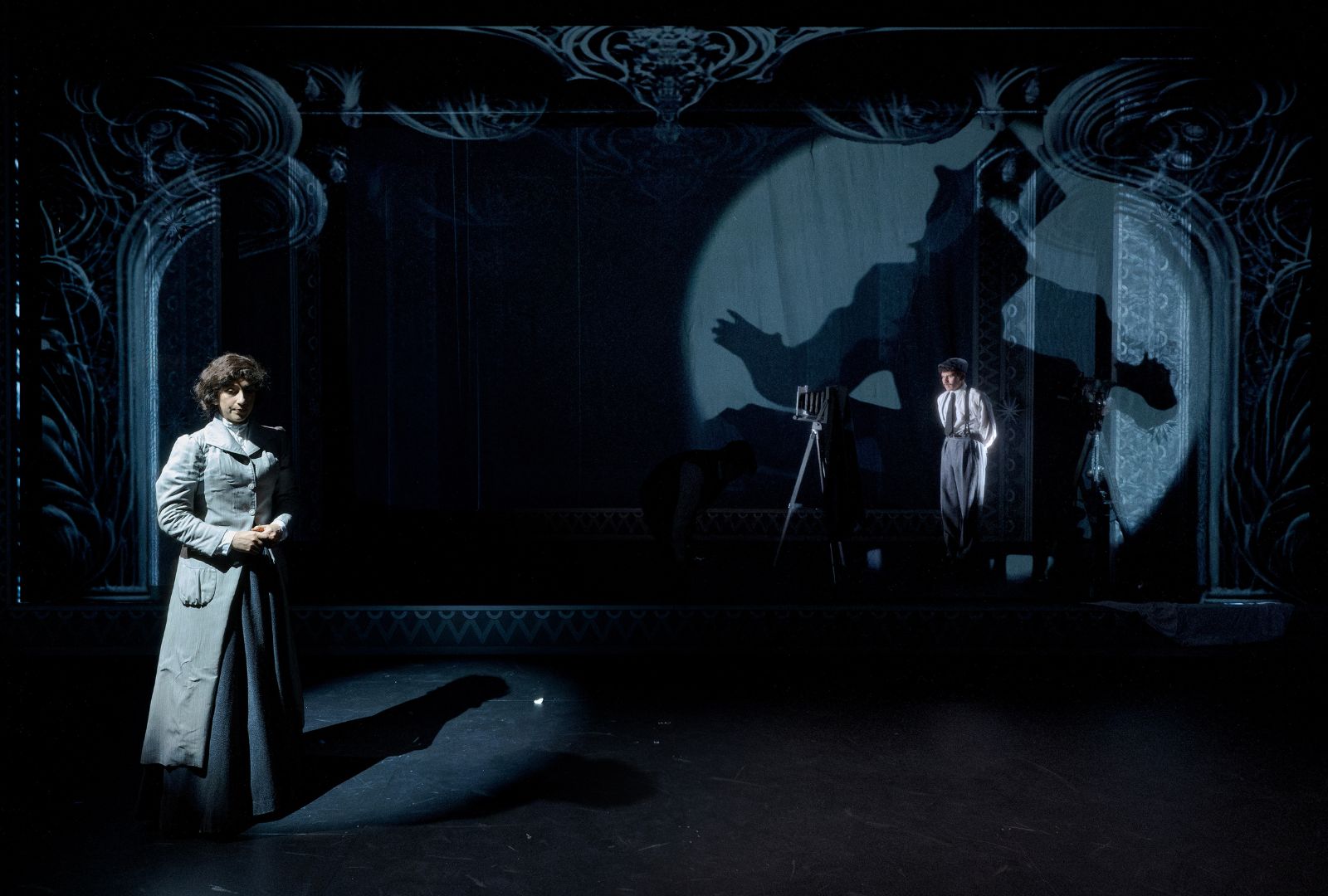A performer in a grey coat on a stage with another performer standing in a spotlight behind them.
