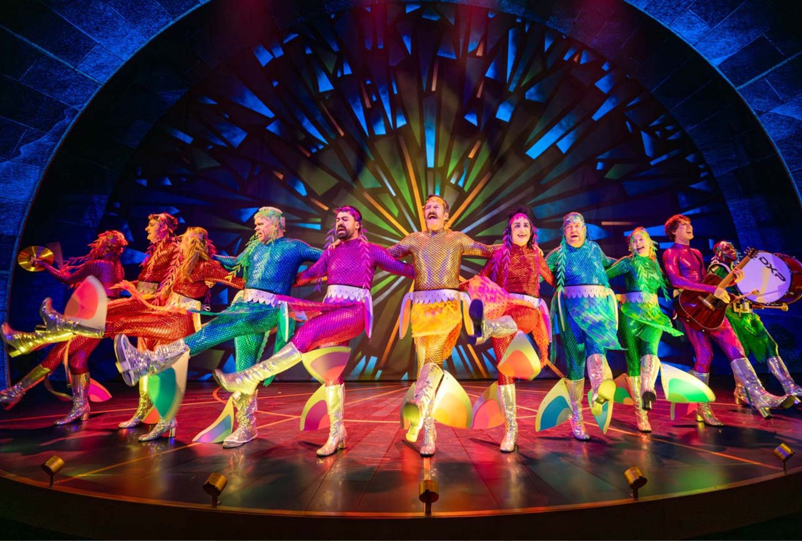 A group of performers in shiny, colourful costumes and silver boots dance in a row on stage, with vibrant rainbow lighting and a dramatic background behind them.