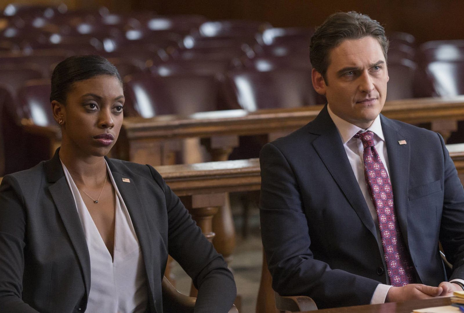 Two actors sitting in a courtroom wearing suits.