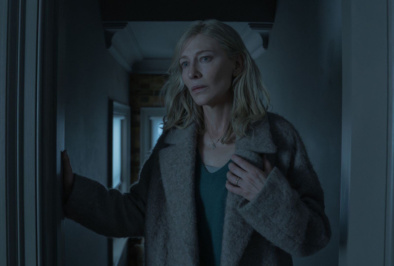 A woman (Cate Blanchett) wearing a coat stands in a hallway, with a hand on the door frame. 
