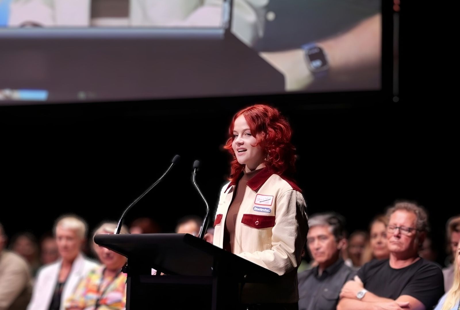 A person on stage presenting to an audience with staff behind them. They have red hair and the podium reads 'NIDA'.