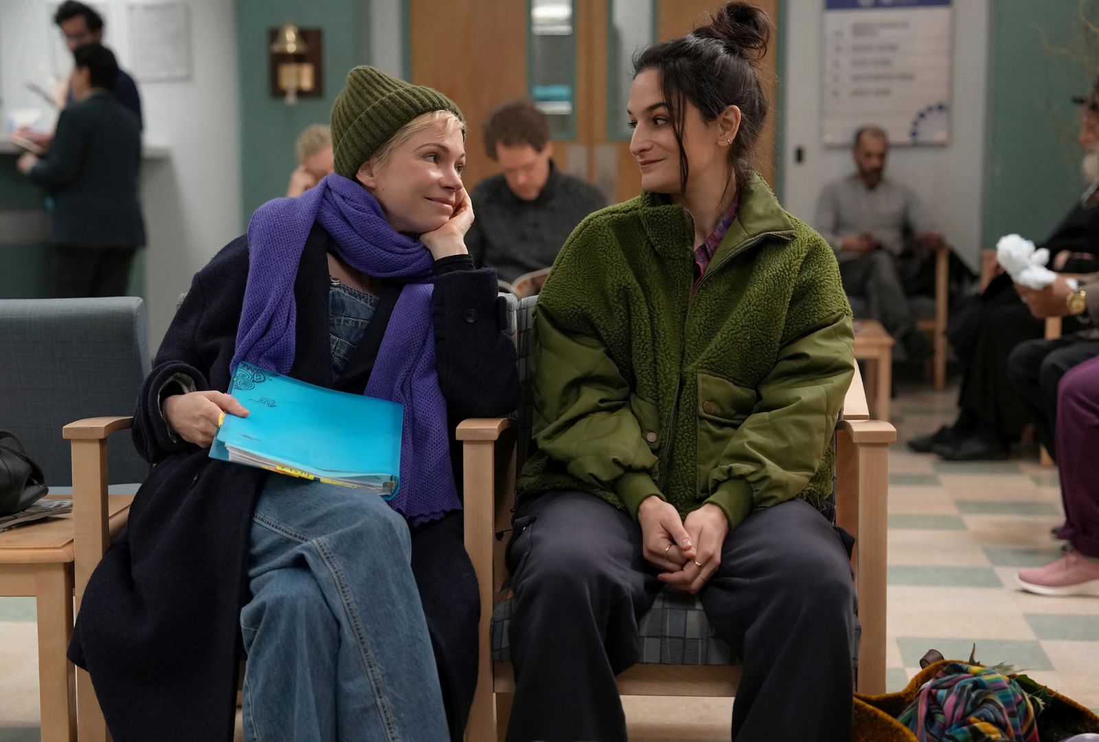 Two women seated in chairs, waiting in a hospital waiting room, appear engaged in conversation.