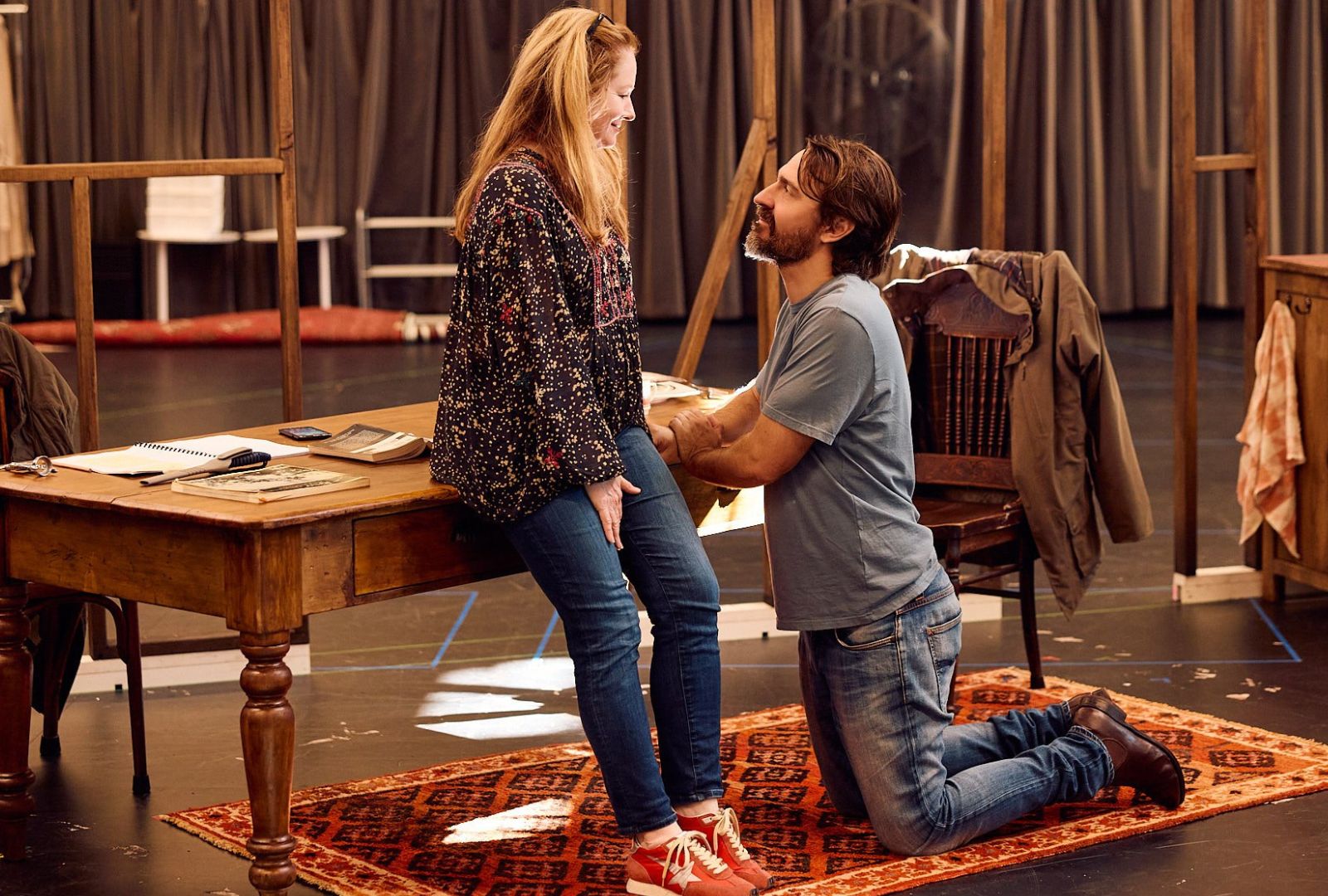 Two people in a rehearsal room, a man in jeans kneels in front of a woman with long hair and red running shoes.