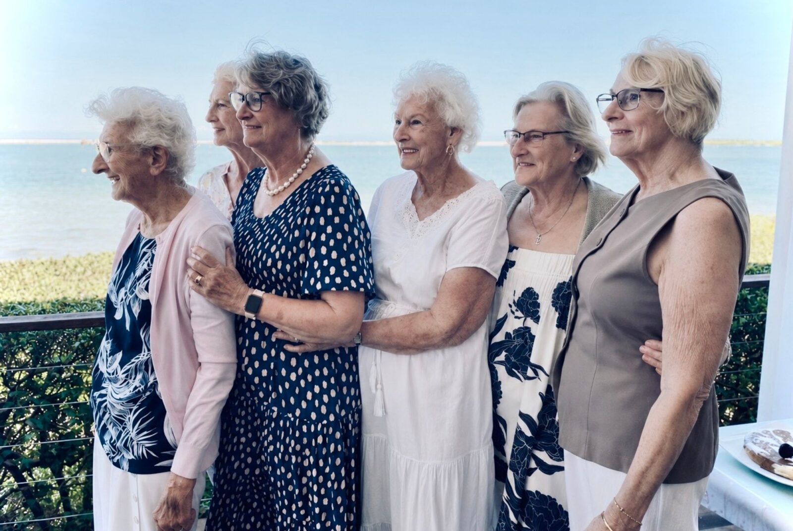 Six women stand together, smiling with their arms around each other. 