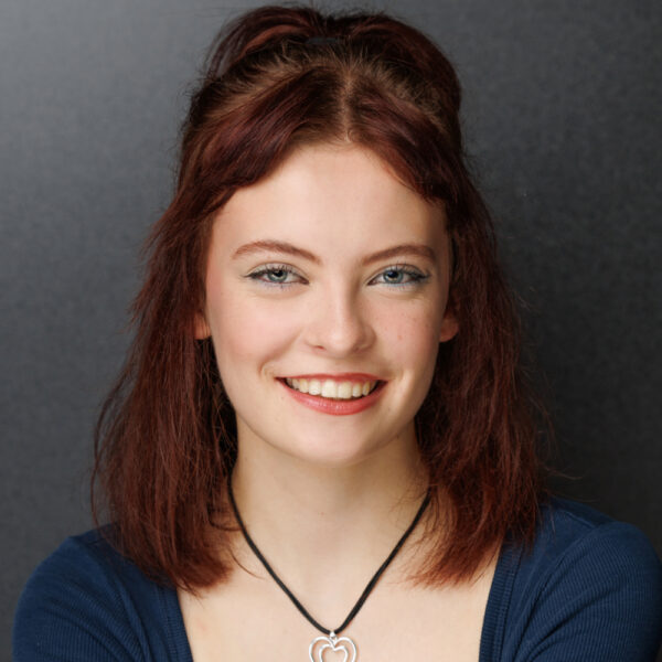 A young woman with shoulder-length auburn hair, blue eyes, and a friendly smile wears a dark blue top and a heart-shaped pendant necklace, posing in front of a plain dark background.