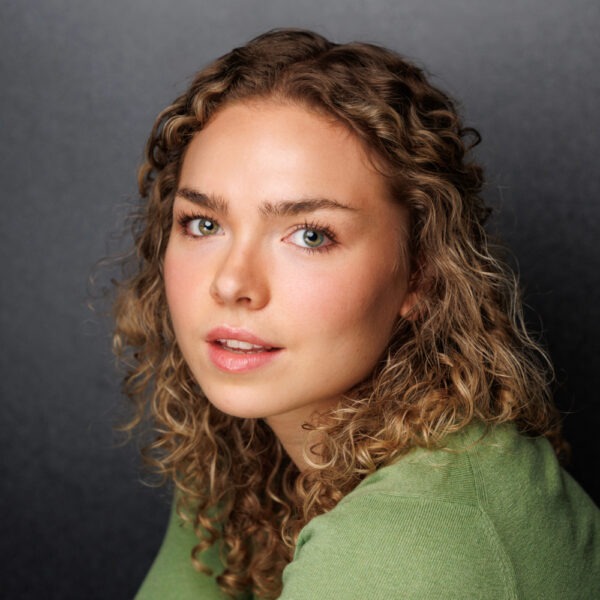 A young woman with curly blonde hair and blue eyes, wearing a light green top, poses in front of a dark grey background, looking softly into the camera with a neutral expression.