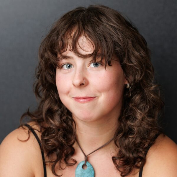A young woman with curly brown hair and blue eyes smiles softly. She has a nose ring, wears a turquoise pendant necklace, and is dressed in a black top against a plain dark background.