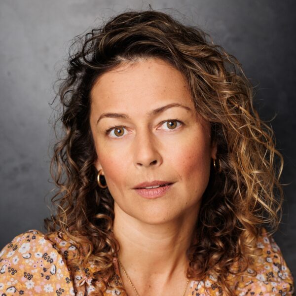 A woman with curly, light brown hair wearing a floral top and gold hoop earrings looks directly at the camera against a dark grey background.