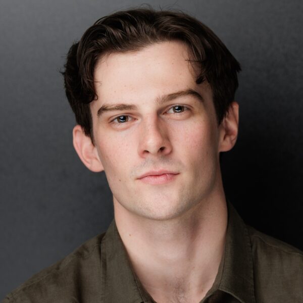 A young man with fair skin, short dark brown hair, and blue eyes looks directly at the camera. He is wearing a dark green collared shirt and is posed against a plain dark background.