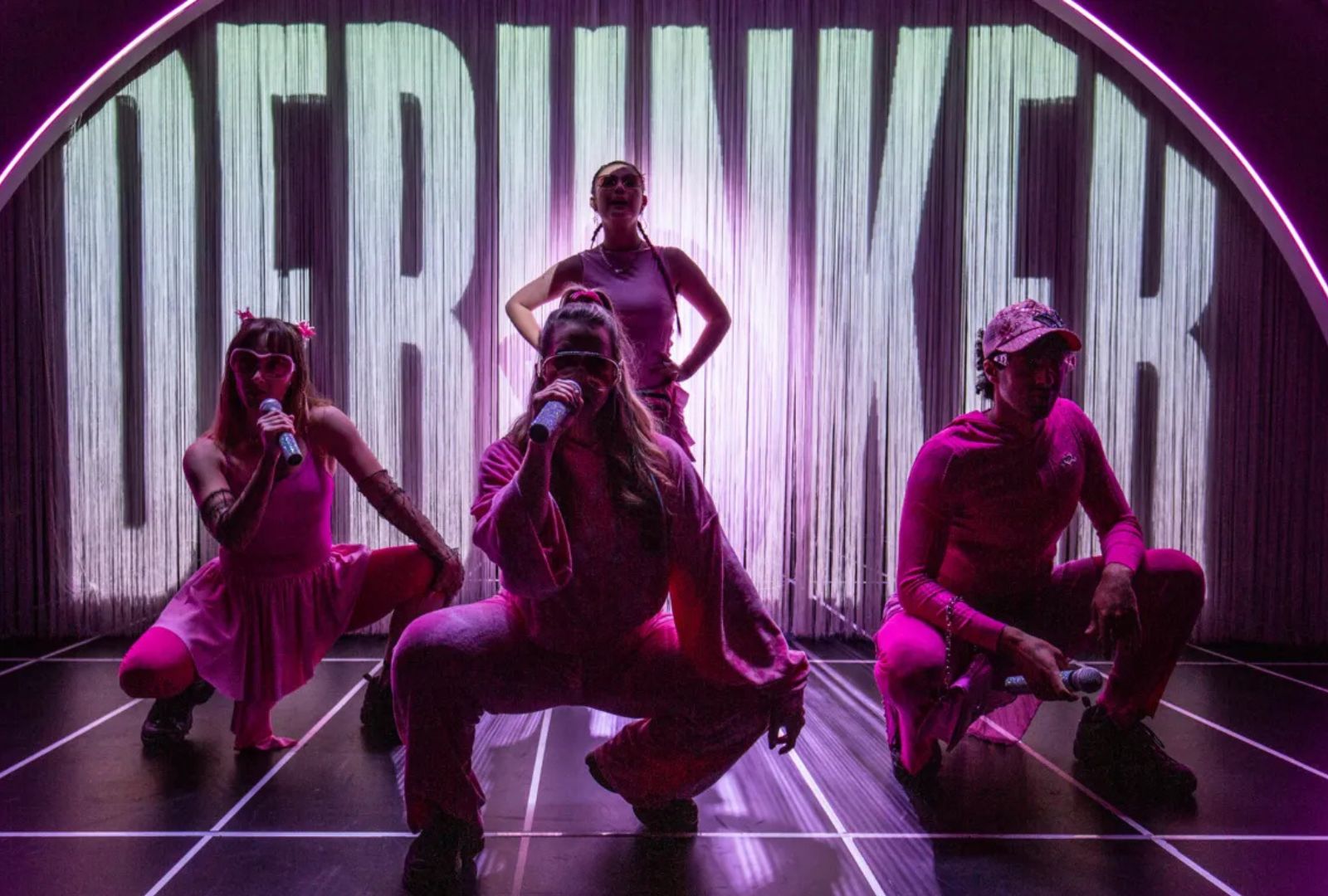 Four performers, three are crouched and singing with one standing behind them. They are in pink light with DEBUNKED projected behind them.