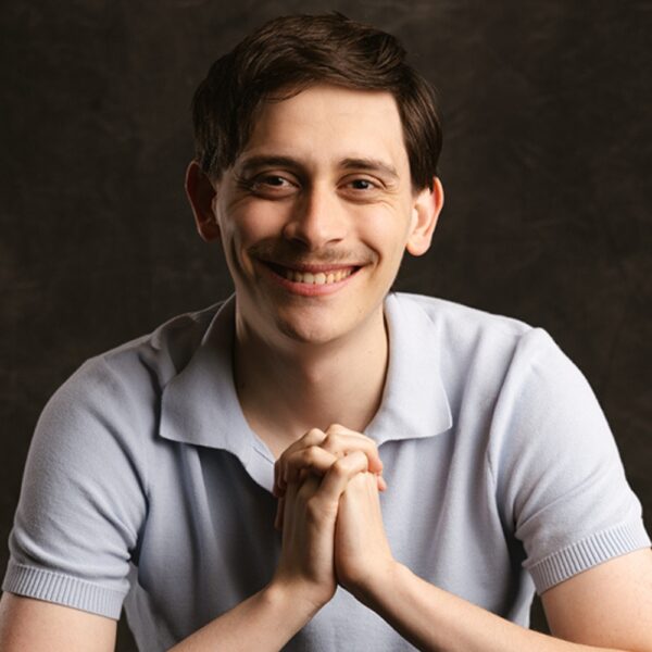 A young man with short brown hair and a light moustache is smiling warmly at the camera, wearing a light blue polo shirt and sitting with his hands clasped together against a dark background.