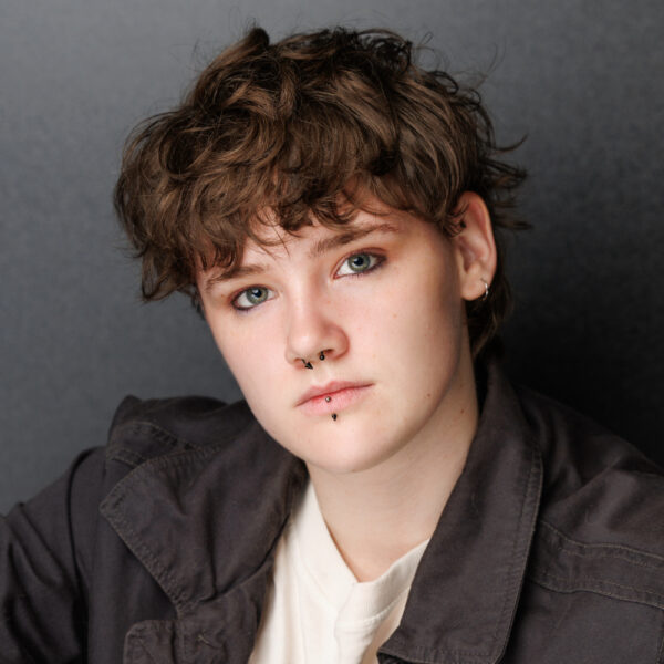 A person with short, tousled brown hair and light eyes looks at the camera. They have a septum piercing, a labret piercing, and wear a dark jacket over a light shirt. The background is plain and dark.