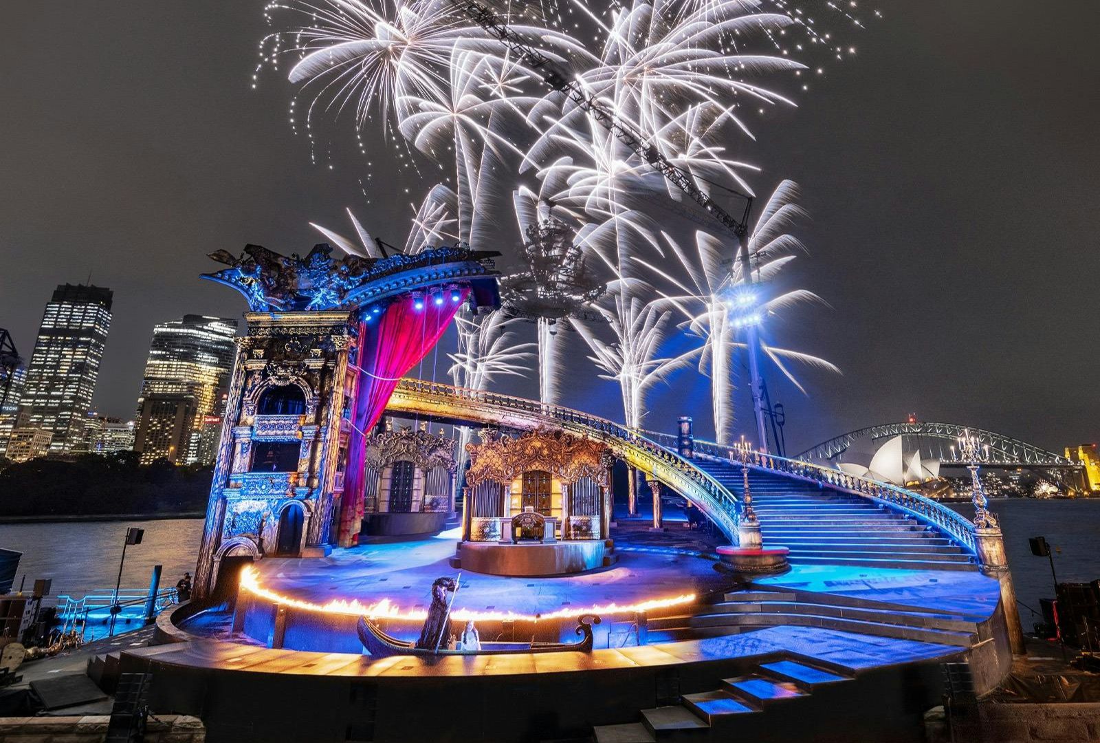 Illuminated opera stage set by the water with ornate architecture and purple lighting. Fireworks explode in the night sky, creating a festive atmosphere.