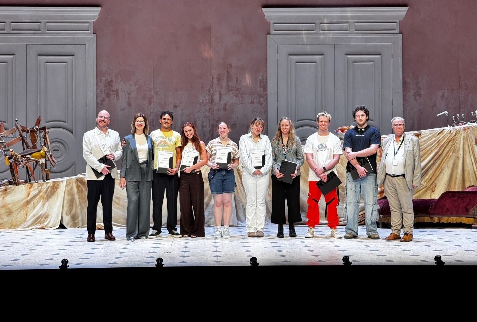 A group of ten people stands in a line on a stage, each holding a certificate. The backdrop features large doors and draped fabric, creating a formal atmosphere. The mood is celebratory and professional.