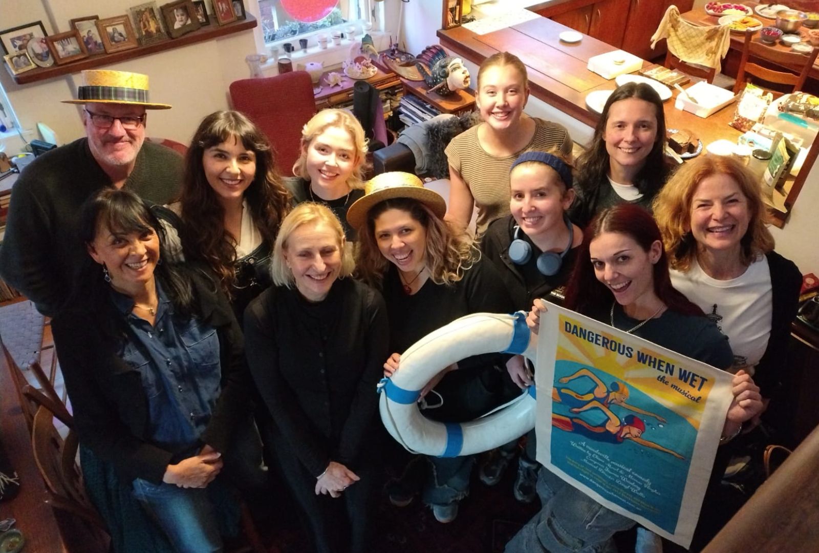 A group of people gathered indoors, some wearing straw hats, smile at the camera. One individual holds a "Dangerous When Wet" poster. The setting appears to be a cozy room with framed pictures and a table laden with items in the background.