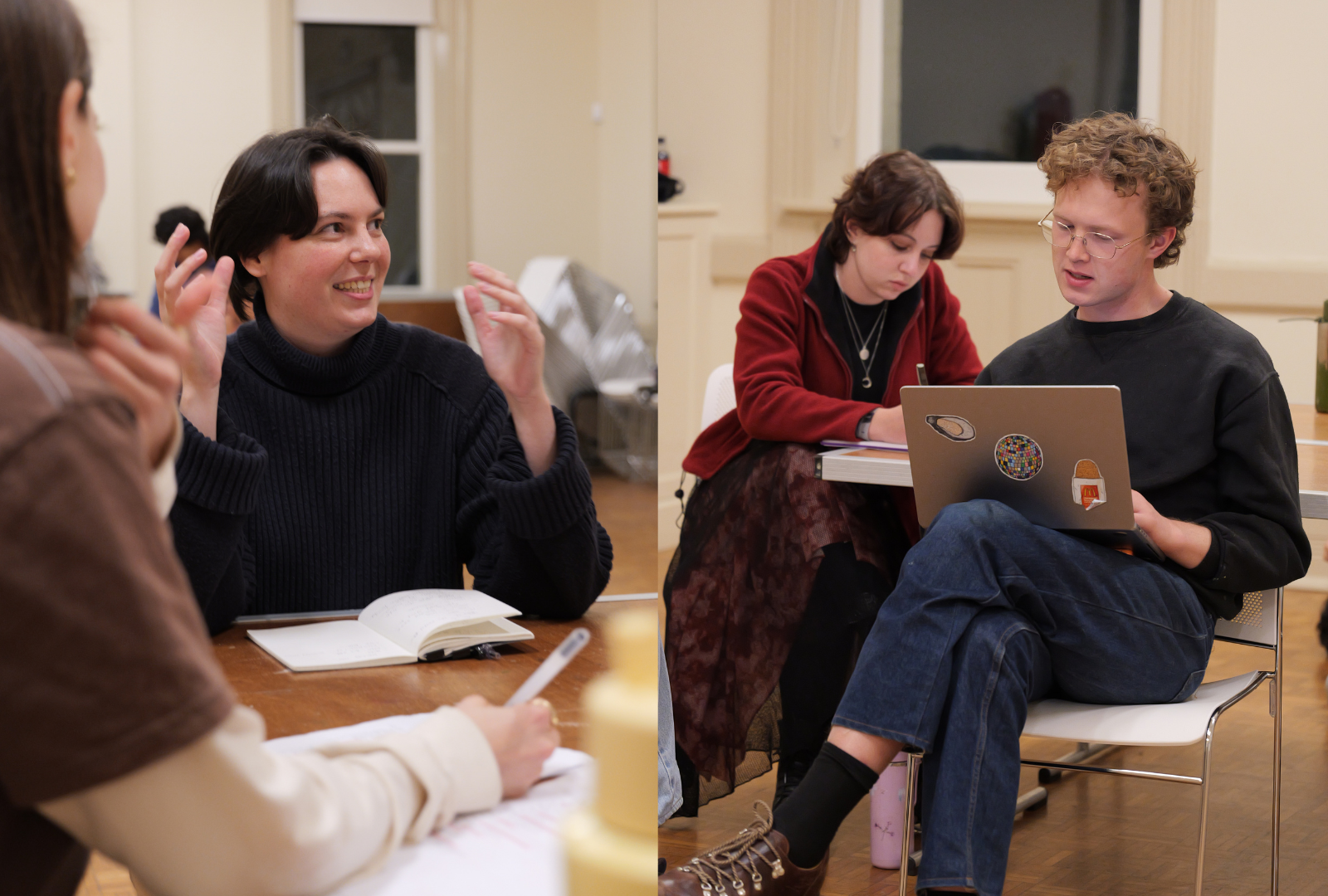 Three individuals are gathered in a room with wooden floors. One person is engaged in conversation, gesturing with their hands while holding a notebook. Two others are seated nearby; one is focused on a laptop with stickers, including a recognizable Microsoft logo. A third person is writing in a notebook, and various items are on the table in front of them.