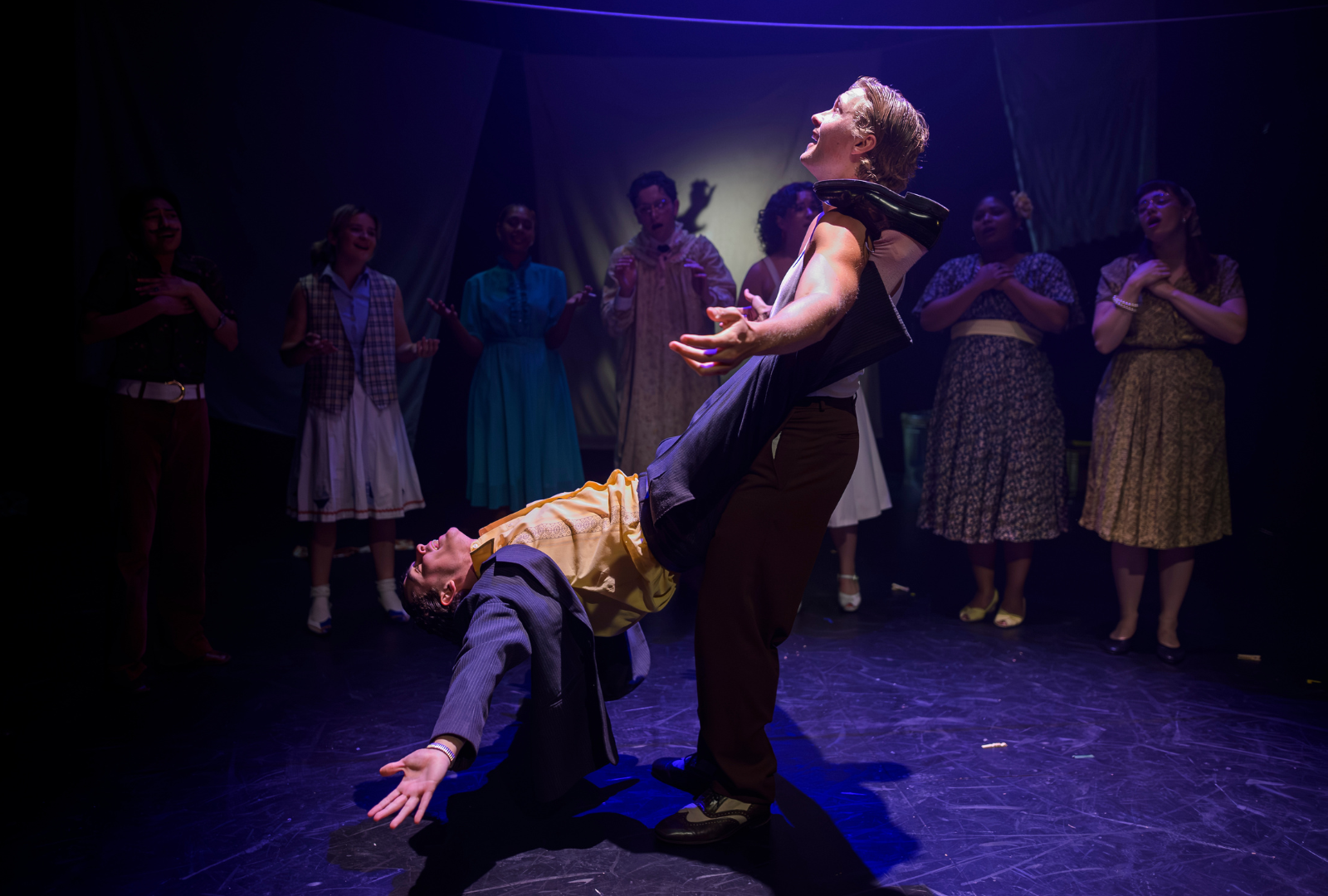 A dynamic stage performance featuring two performers in the foreground. One performer is leaning back dramatically, supported by another. Several other performers, including recognizable figures, stand in the background, observing and clapping. The scene is colorful and illuminated by stage lighting, creating a lively atmosphere.