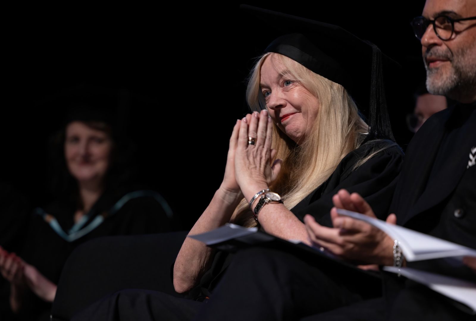Suzie Miller sits with her hands together in a graduation gown looking towards the speaker. 