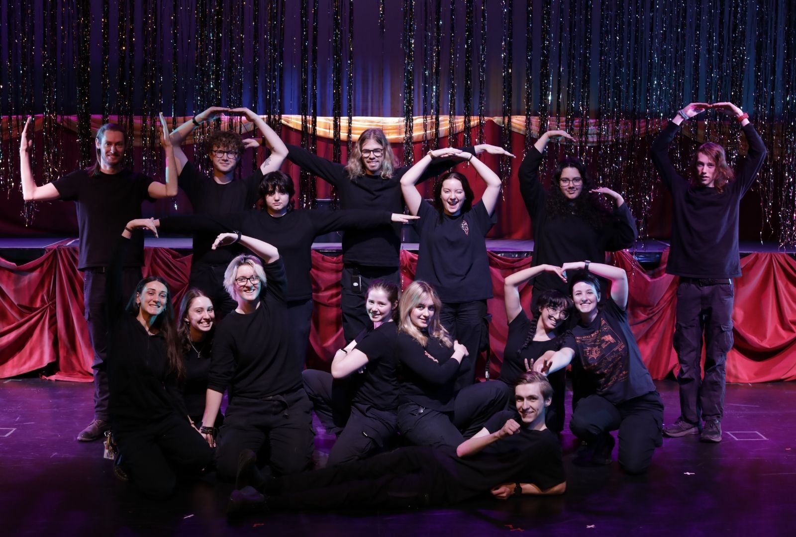 A group of individuals dressed in black pose joyfully on stage under a shimmering backdrop, forming different shapes with their arms. The setting includes sparkling vertical strands and red drapes.