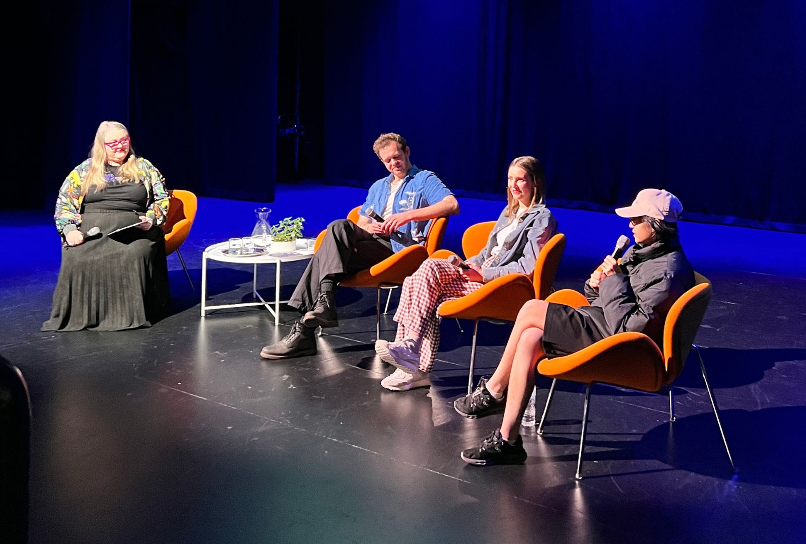A panel of four individuals sits on stage in orange chairs, engaged in discussion. The setting is a dimly lit auditorium with a blue backdrop. A round table with a plant and water glasses is positioned in front of them.