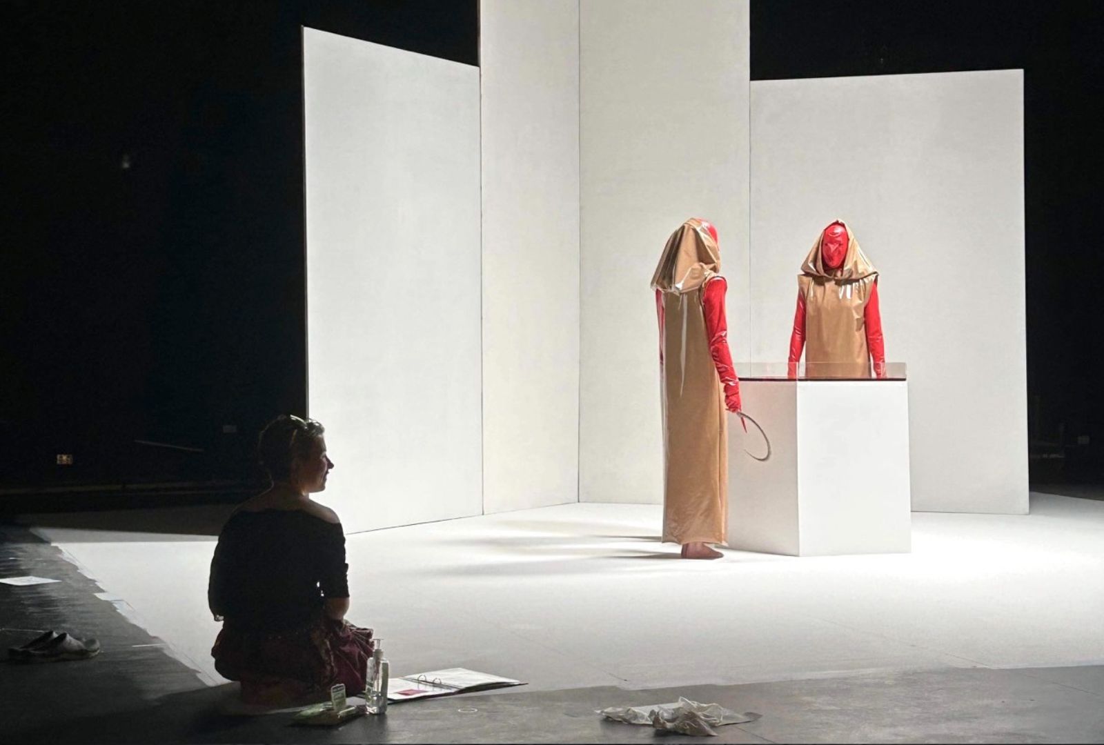From left to right: Paris Burrows sits on the ground watching two actors (right) dressed in red masks and beige cloaks on a white set. The hold sickles.