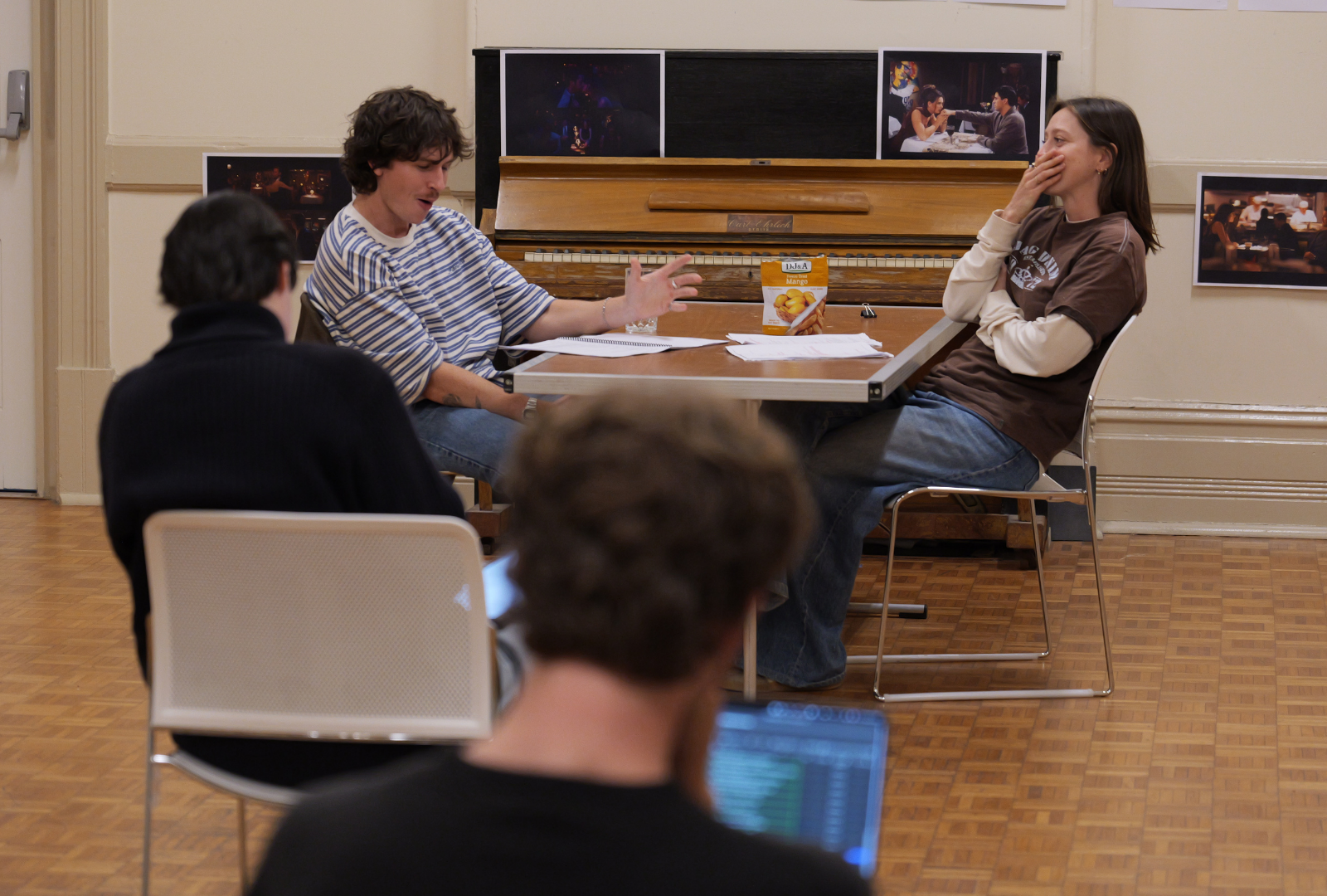 People are seated around a table in a room with a piano and musical posters on the wall. Two individuals are engaged in a conversation at the table with papers and colourful snacks between them. Two others are seated with their backs to the camera, one using a laptop.