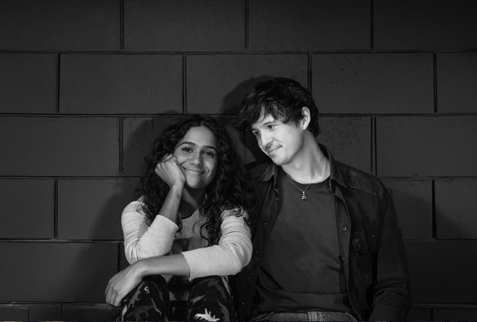Alyssa Peters (left) and Josh Hammond (right) sit on the ground in front of a brick wall smiling. The photo is in black and white.