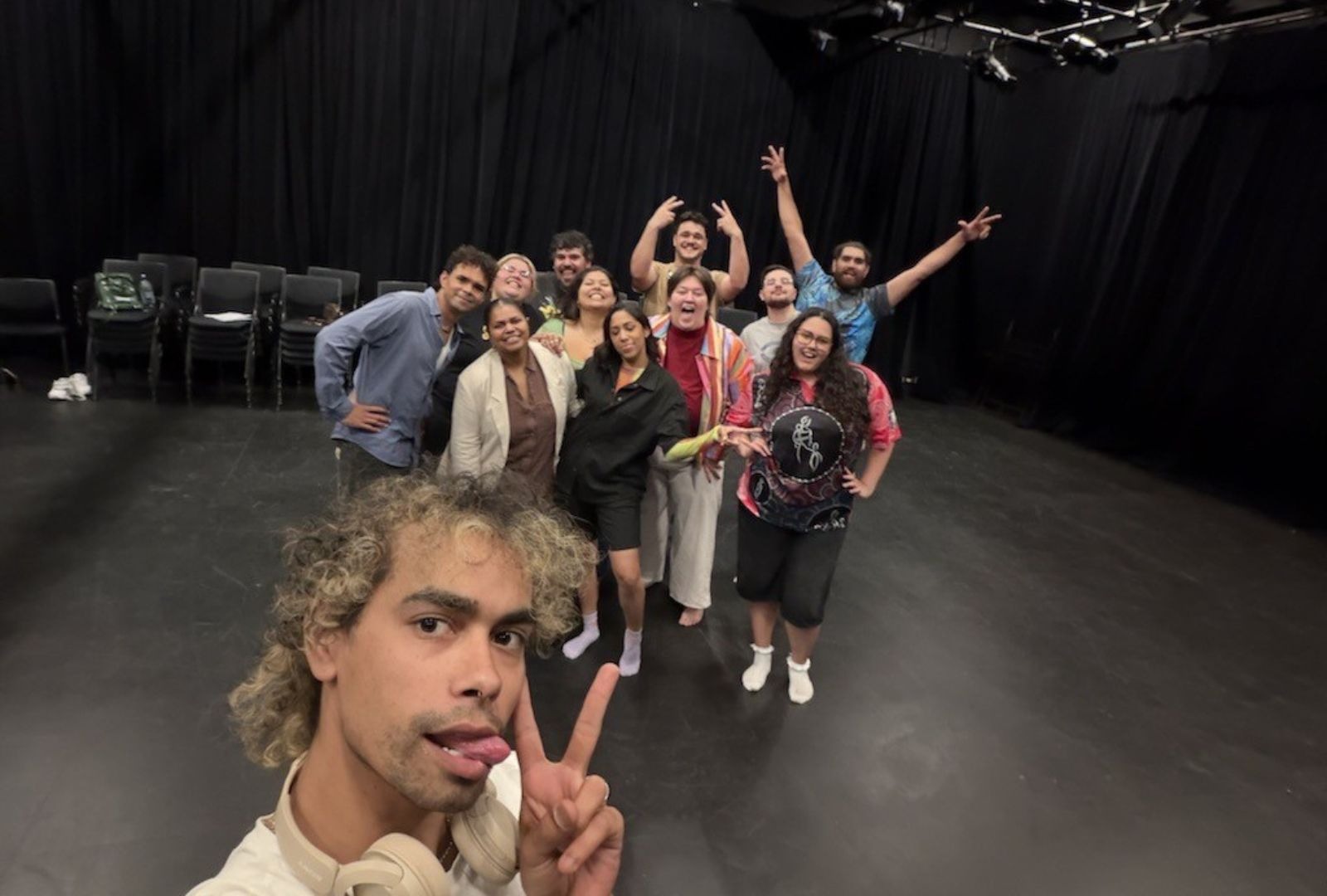 A group of people poses together on a stage with black curtains and empty chairs in the background. The person in front takes a selfie, making a peace sign and sticking their tongue out.