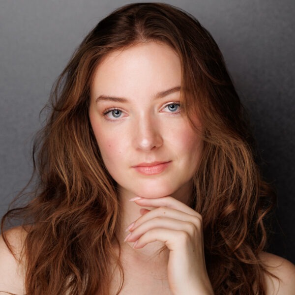 A young woman with long, wavy brown hair and blue eyes poses in front of a grey background, gently touching her chin with her fingers. She has a natural expression and minimal make-up.
