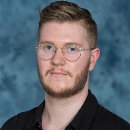 A young man with short, light brown hair, a trimmed beard, and round glasses, wearing a black shirt, stands in front of a blue, mottled background.