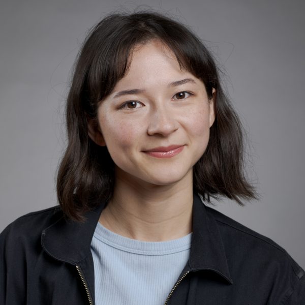 A young woman with shoulder-length dark hair and fringe smiles softly. She is wearing a light blue top and a dark jacket, posing in front of a plain grey background.