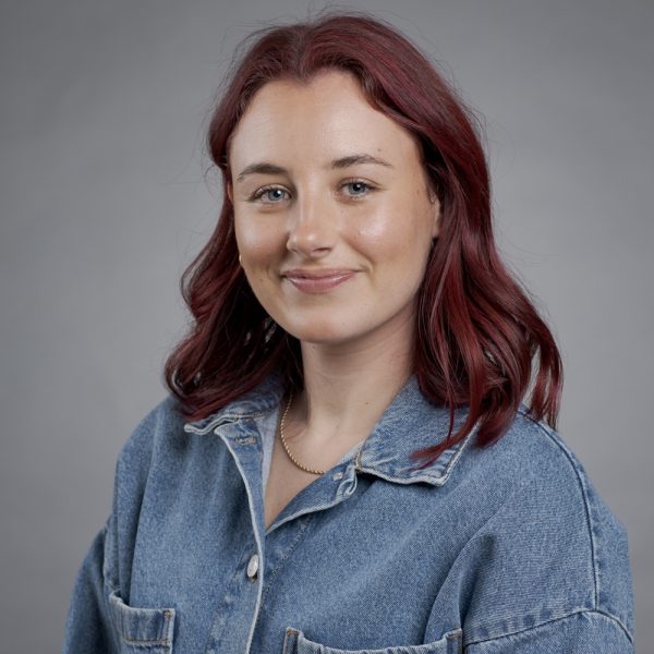 A woman with shoulder-length red hair wearing a denim jacket and a gold necklace smiles softly at the camera against a plain grey background.