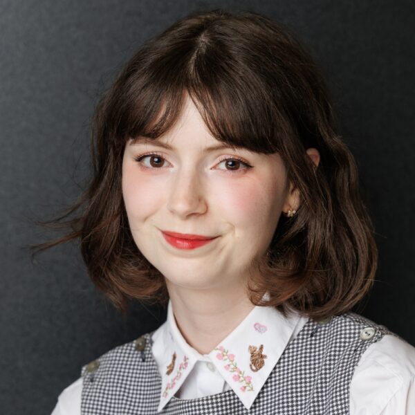 A young woman with short brown hair and a fringe smiles softly. She wears a white collared shirt with floral embroidery and a black-and-white chequered dress, posed against a dark grey background.