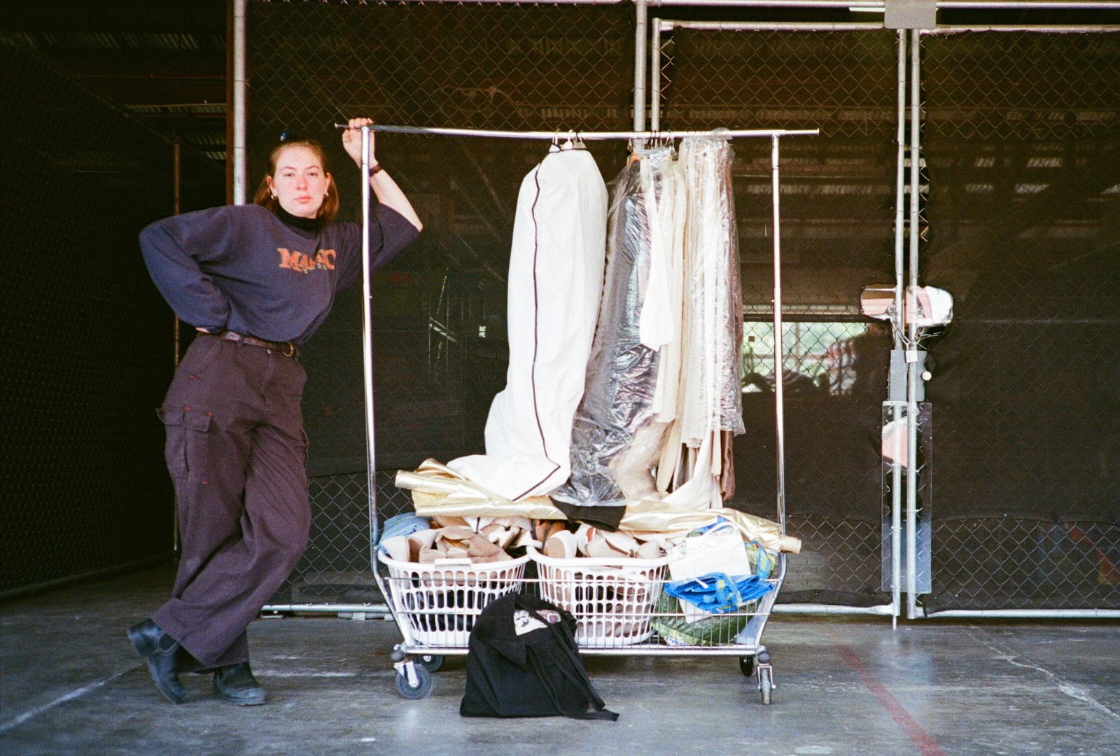 A person leans on a rolling clothes rail filled with clothes and fabric rolls in a large industrial setting, next to a black bag on the ground. The background has metal fencing and a partially open gate.