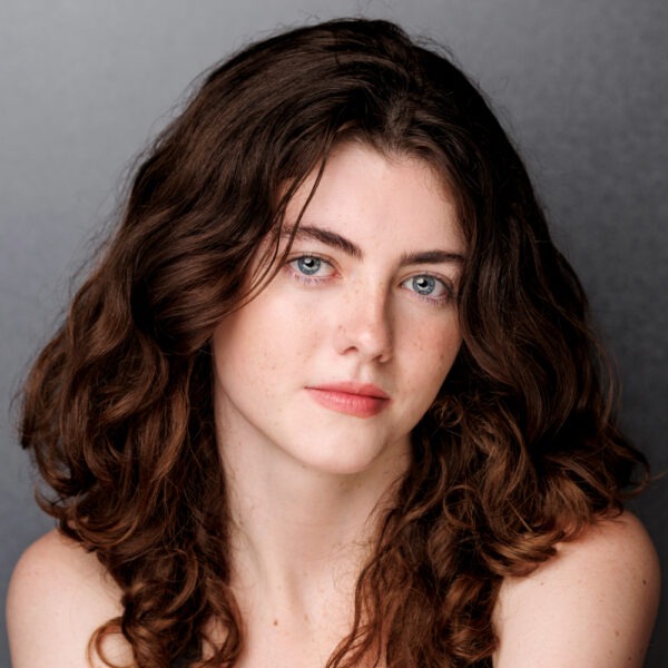 A young woman with long, curly brown hair and blue eyes looks at the camera with a neutral expression, posing against a plain dark grey background.