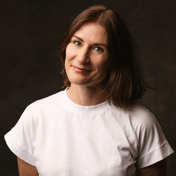 A woman with shoulder-length brown hair smiles gently whilst wearing a plain white T-shirt, standing against a dark, neutral background.